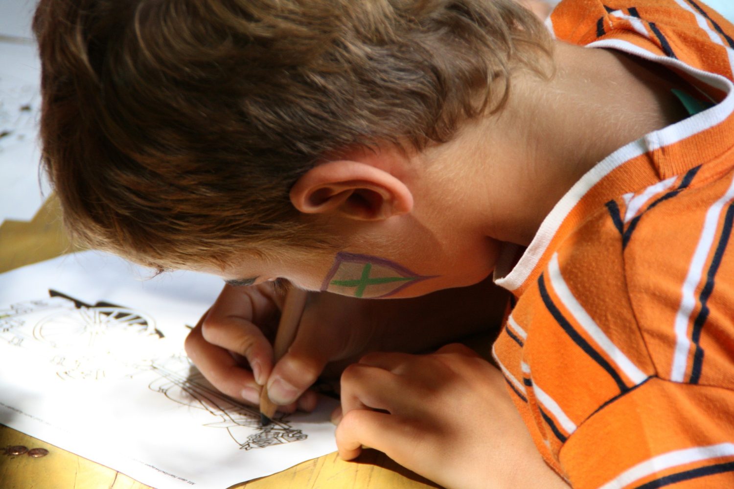 Close up of side of head and hands of child drawing on a piece of paper on top of a school desk