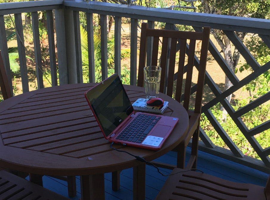 Bahamian hotel balcony, with wooden fence, table and chairs. Laptop on table.