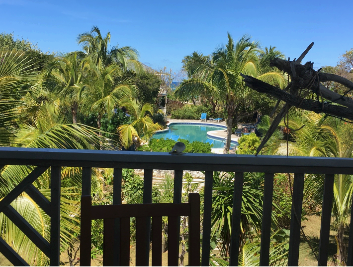 View of pool from Bahamian hotel wooden balcony. Blue pool, lush green palms and vegetation, ocean beyond