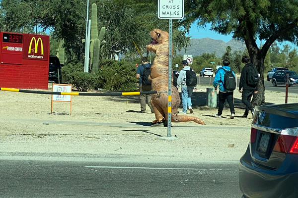 A person in a t-rex costume on the sidewalk waiting to cross a street.