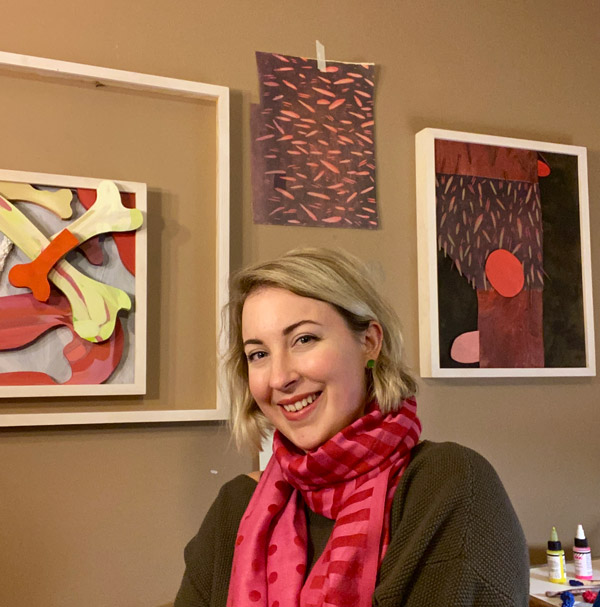 Woman with short blonde hair and a red scarf smiling, her artwork is hanging on a wall behind her.