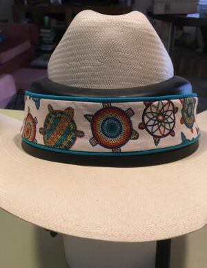 A light colored cowboy hat with a protective layer around the the crown. A turtle embroidered fabric band is wrapped around it.