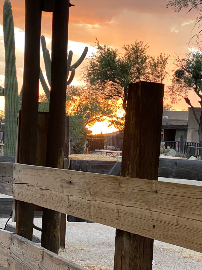 A bright orange sunset glows above the wooden fencing of a corral.